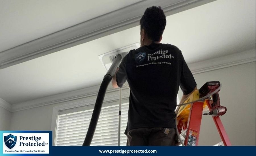 An air duct cleaning technician standing on a ladder to vacuum a ceiling vent using professional equipment.